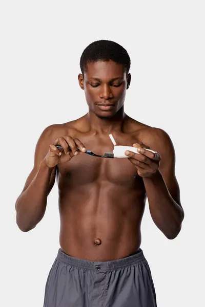 stock image A shirtless African American man holds a toothbrush and toothpaste, ready for his beauty routine in a studio with a white background.