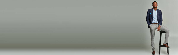 A handsome African American man in formal attire, wearing a checkered blazer, sitting confidently on top of a stool.