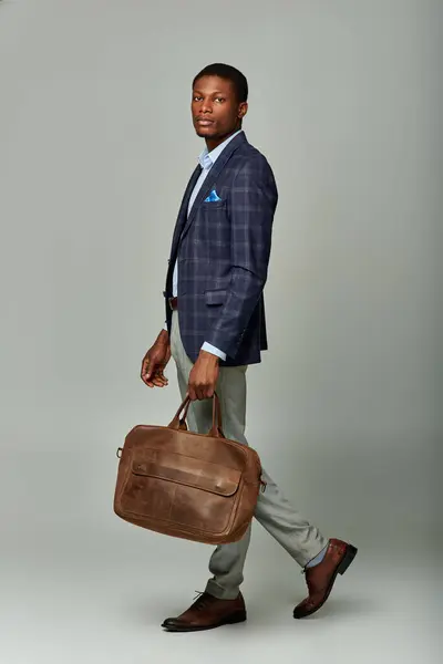 stock image Handsome African American businessman in checkered blazer carrying a brown bag on grey background.