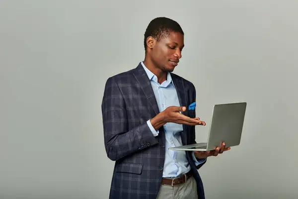 stock image Young African American businessman in checkered blazer operating laptop against grey backdrop.