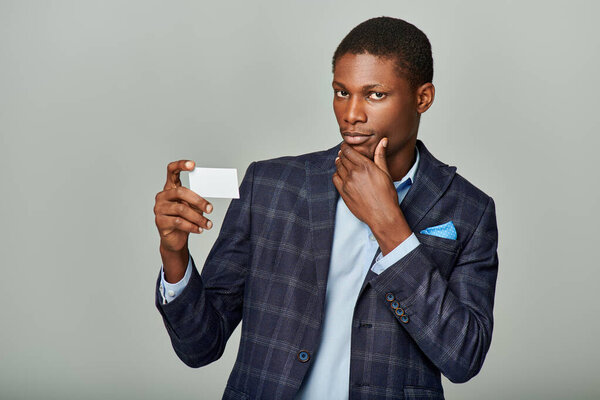 Афроамериканский бизнесмен в клетчатом пиджаке представляет визитку на сером фоне.