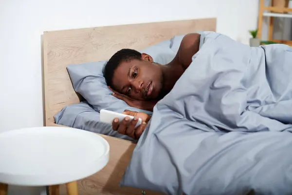 stock image A young African American man lays comfortably in bed, holding a remote control in his hand as he wakes up.