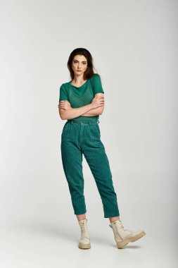 Zümrüt yeşili kadife pantolon ve yeşil gömlek giyen genç bir kadın kollarını kavuşturmuş poz veriyor..