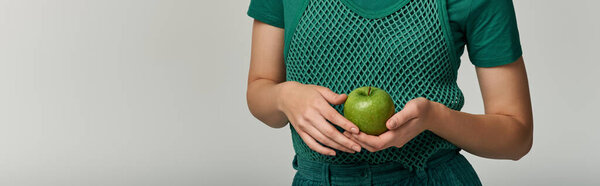 A woman in a green top and trousers holds a green apple in her hands.