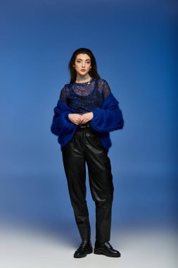 Siyah deri pantolonlu ve mavi tüylü hırkalı genç bir kadın mavi bir arkaplanda duruyor..