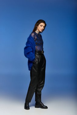 Mavi hırka ve siyah pantolon giyen genç bir kadın mavi bir fonda poz veriyor..