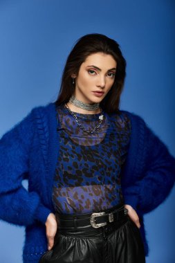 Mavi hırkalı ve leopar desenli bluzlu genç bir kadın parlak mavi bir arka planda duruyor..