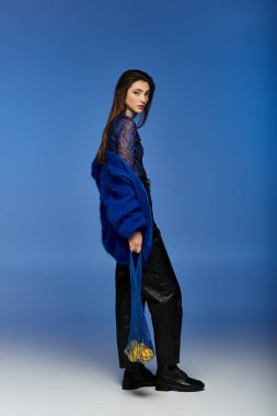 Mavi ceketli genç bir kadın elinde meyve dolu bir çantayla mavi bir arkaplanda duruyor..