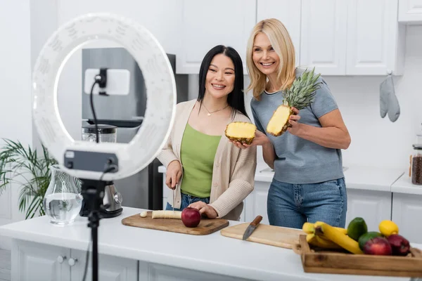 Cheerful interracial friends cutting fruits near blender and smartphone in kitchen — Stock Photo