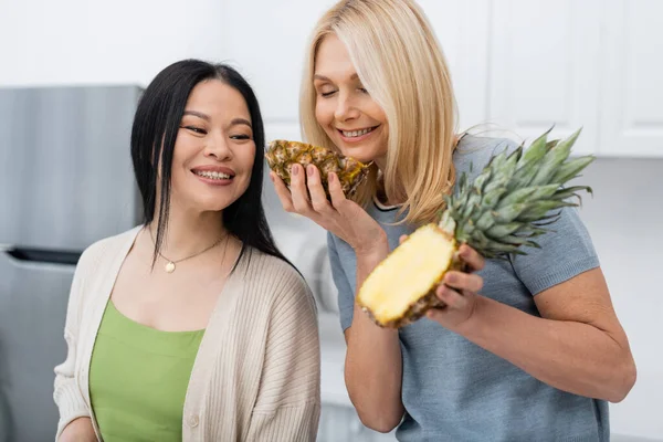 Smiling woman smelling fresh pineapple near asian friend in kitchen — Stock Photo