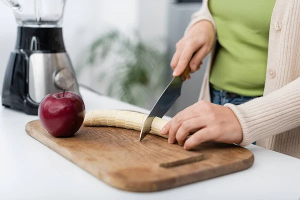 Cropped view of woman cutting banana near apple and blurred blender in kitchen — Stock Photo