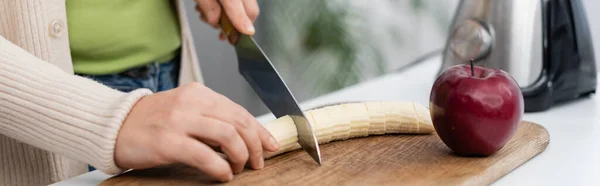 Vista cortada da mulher cortando banana fresca perto de maçã e liquidificador na cozinha, banner — Fotografia de Stock