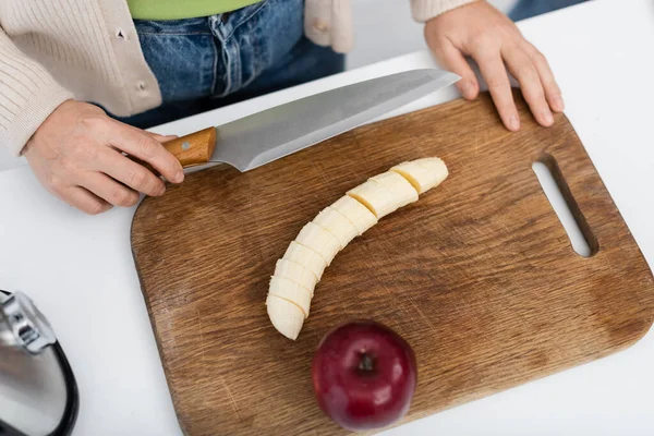Top view of woman holding knife near banana and apple on cutting board — Stock Photo