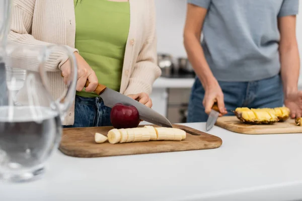 Cropped view of woman cutting fresh apple near banana and blurred friend in kitchen — Stock Photo
