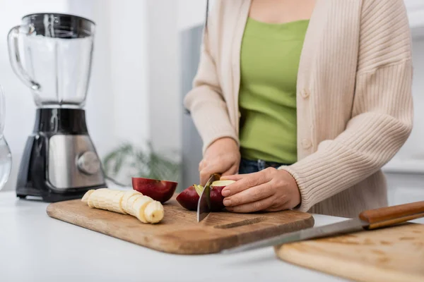 Cropped view of woman cutting ripe apple near banana and blurred blender in kitchen — Stock Photo