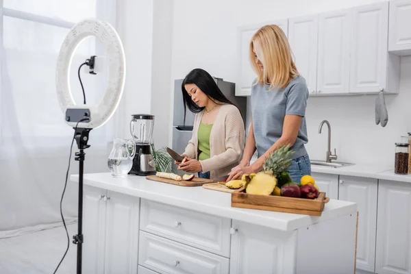 Міжрасові блогери ріжуть свіжі фрукти біля блендера і смартфона на кухні — стокове фото