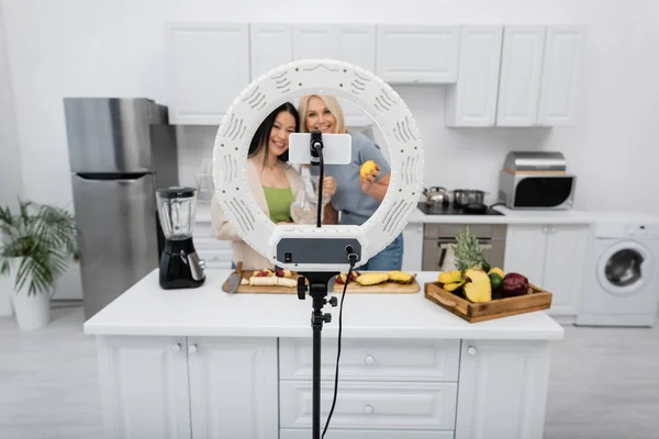Smartphone and ring light near blurred multiethnic friends with fruits in kitchen — Stock Photo