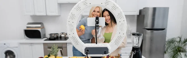Smartphone y anillo de luz cerca borrosa amigos multiétnicos con frutas frescas en la cocina, pancarta - foto de stock