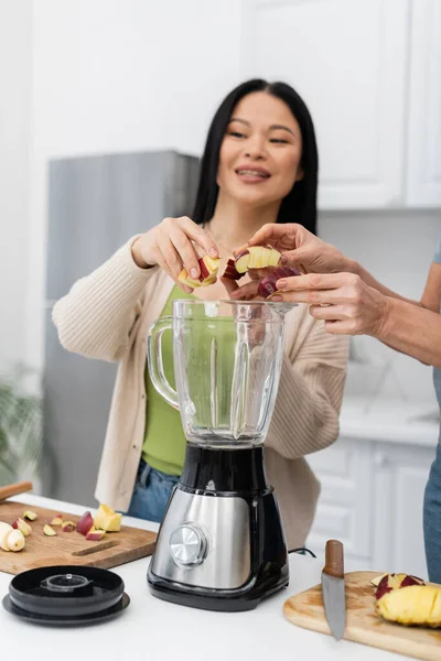 Woman putting apple in blender near blurred asian friend in kitchen — Stock Photo