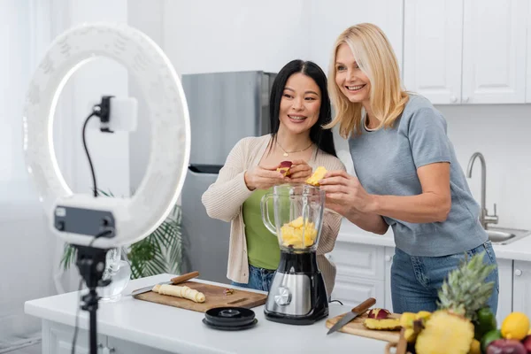 Positive multiethnic women putting fruits in blender near smartphone and ring light in kitchen — Stock Photo