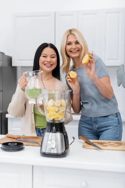 Positive interracial friends holding fruits and water near blender in kitchen — Stock Photo