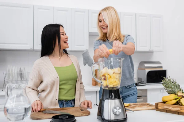 Cheerful multiethnic friends talking while preparing smoothie in kitchen — Stock Photo