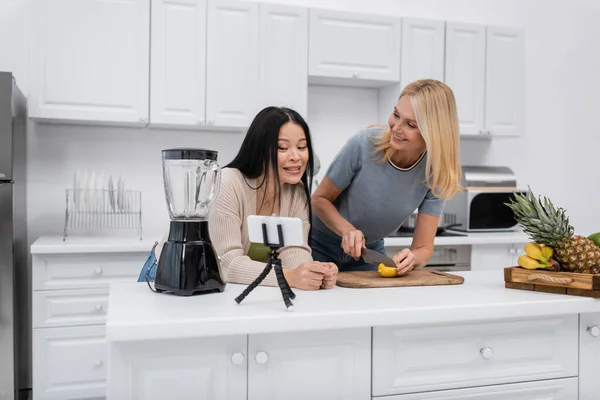 Усміхнена азіатська жінка говорить біля смартфона на тринозі, а друг ріже фрукти на кухні — стокове фото