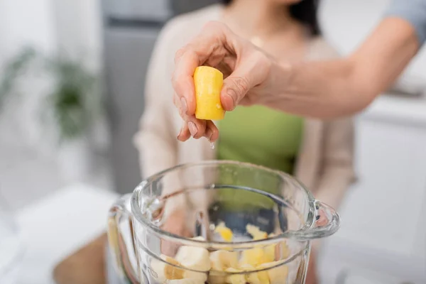 Cropped view of woman squeezing lemon above blender with fruits in kitchen — Stock Photo