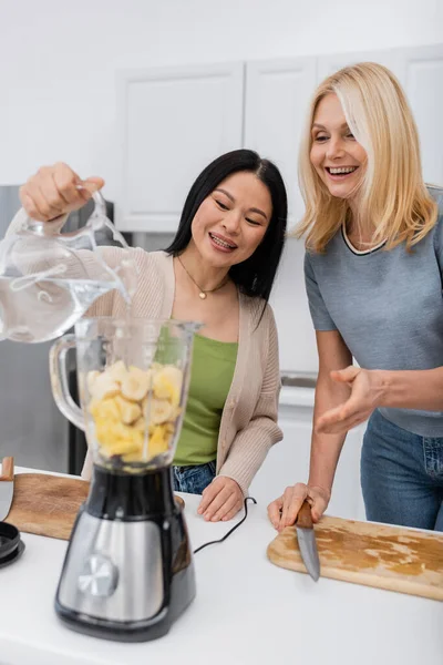 Asiatico donna versando acqua in frullatore con frutta vicino amico in cucina — Foto stock