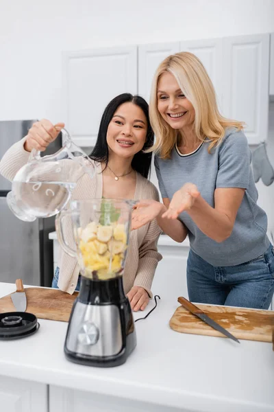Sourire femme pointant avec les mains près de l'ami asiatique verser de l'eau dans le mélangeur avec des fruits dans la cuisine — Photo de stock