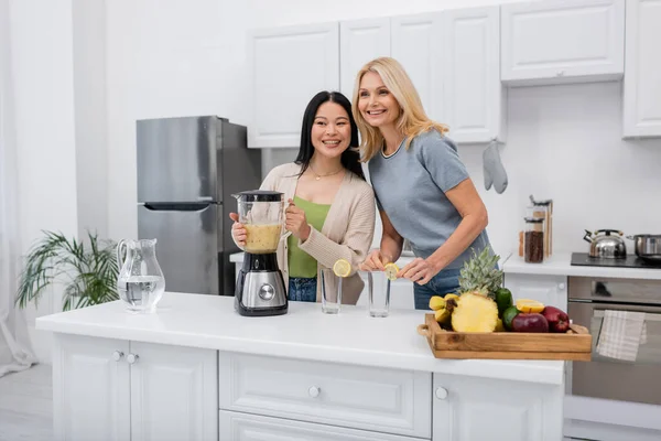 Smiling interracial women holding blender with fruit smoothie near glasses in kitchen — Stock Photo