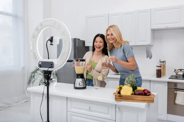 Fröhliche multiethnische Frauen halten Smoothie und Glas neben Smartphone mit Ringlampe in der Küche — Stockfoto