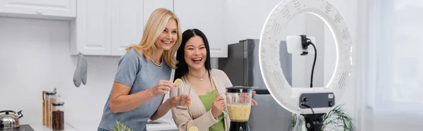 Multiethnic bloggers holding glass and blender with smoothie near smartphone and ring light at home, banner — Stock Photo