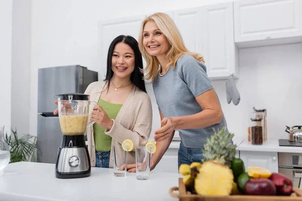 Cheerful interracial women looking away near smoothie and glasses in kitchen — Stock Photo