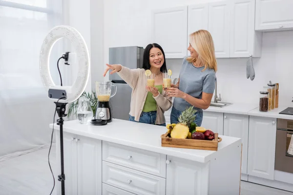 Smiling asian blogger pointing at smartphone and ring light near friend with smoothie in kitchen — Stock Photo