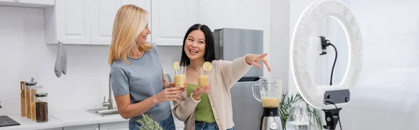 Cheerful asian blogger holding smoothie and pointing at smartphone in ring light near friend at home, banner — Stock Photo