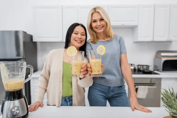 Blurred interracial friends smiling and looking at camera while holding smoothie in kitchen — Stock Photo