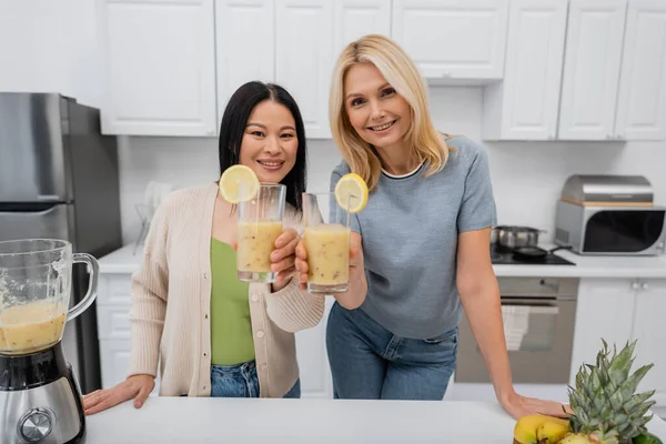 Smiling interracial friends looking at camera and holding glasses of fruit smoothie in kitchen — Stock Photo