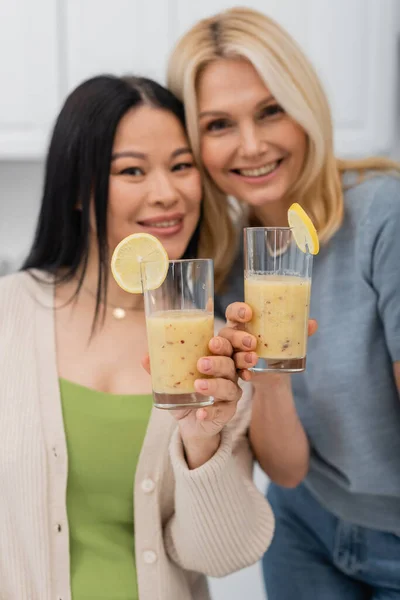 Blurred interracial friends looking at camera and holding fruit smoothie at home — Stock Photo