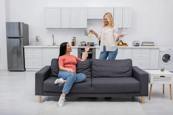 Smiling woman holding champagne bottle and glasses near asian friend at home — Stock Photo