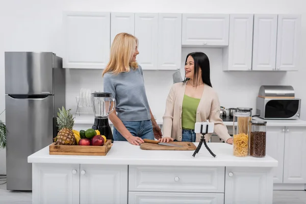 Positive interracial women standing near food and smartphone on tripod at home — Stock Photo
