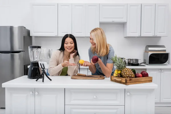 Femmes multiethniques positives pointant du doigt les fruits près du smartphone sur le trépied dans la cuisine — Photo de stock