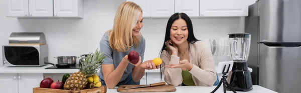 Smiling asian woman pointing at fruits near friend and smartphone on tripod in kitchen, banner — Stock Photo