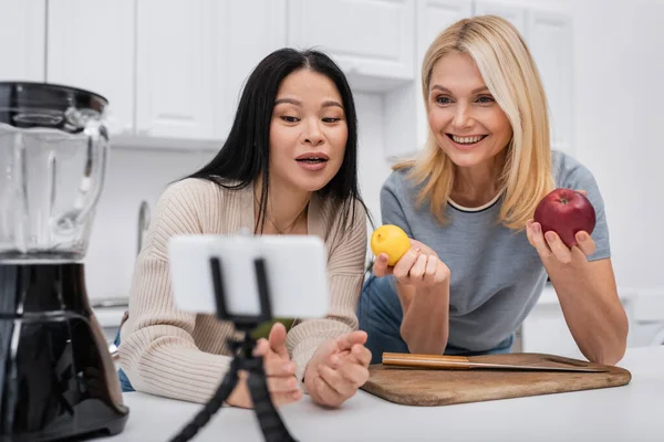 Alegre mujer sosteniendo frutas mientras asiático amigo hablando en smartphone en trípode en cocina - foto de stock