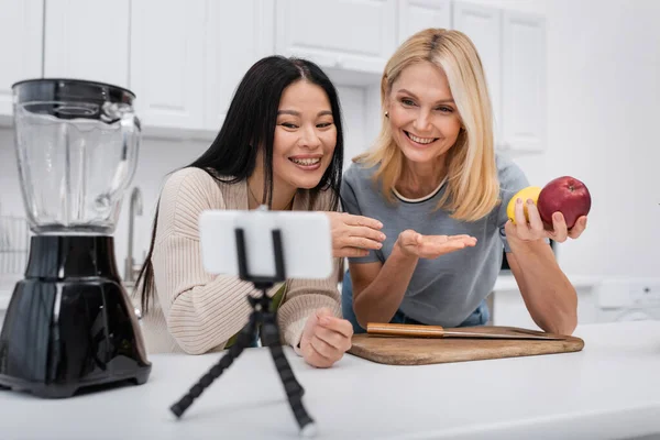 Positive interracial women pointing at fruits near blender and smartphone on tripod in kitchen — Stock Photo