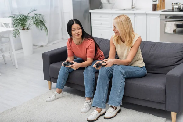 KYIV, UCRANIA - 2 DE DICIEMBRE DE 2021: Mujeres multiétnicas jugando videojuegos y mirándose en el sofá en casa - foto de stock