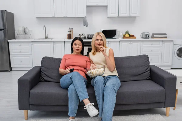 Interracial amis tenant pop-corn tout en regardant la télévision sur le canapé à la maison — Photo de stock
