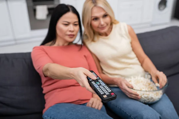 Mujeres multiétnicas borrosas sosteniendo palomitas de maíz y haciendo clic en canales en casa - foto de stock