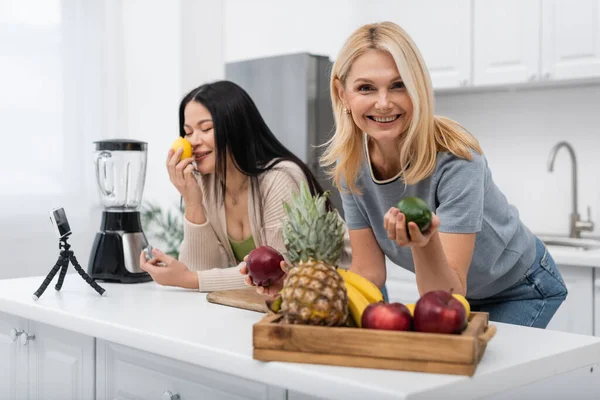 Sonriente mujer sosteniendo fruta cerca asiático amigo y smartphone en trípode en cocina - foto de stock