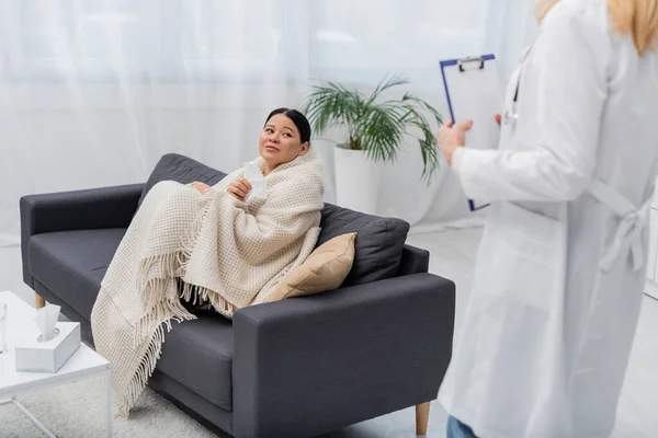 Sick asian woman holding napkin near blurred doctor with clipboard at home — Stock Photo